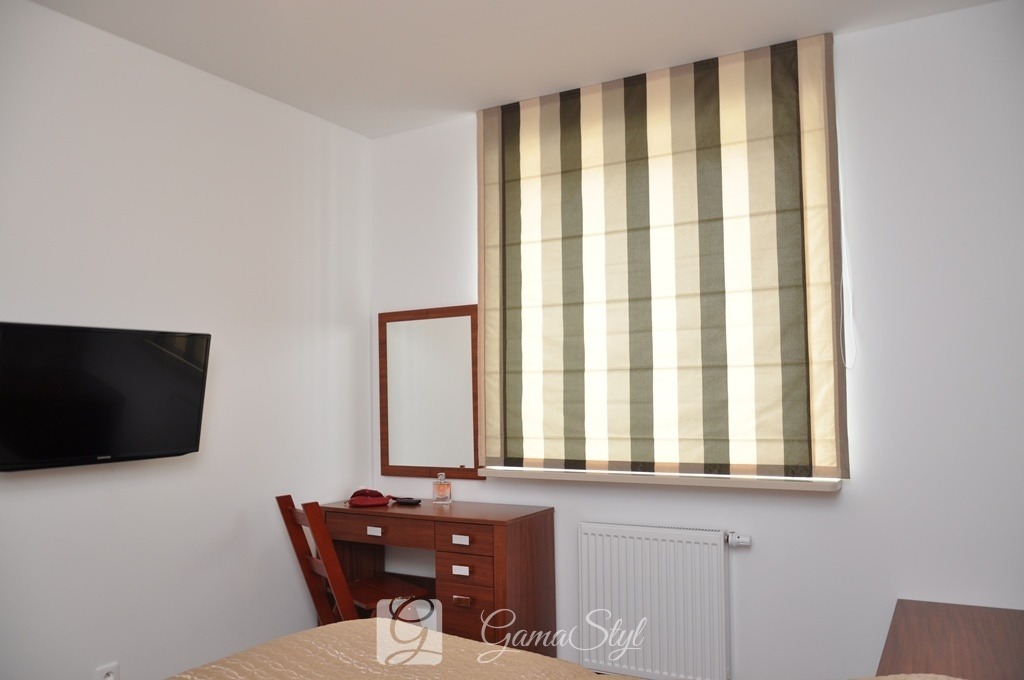 Wnętrze pokoju z roletą rzymską w szerokie pasy w odcieniach beżu i szarości, wiszącą w oknie nad biurkiem z lustrem i grzejnikiem.