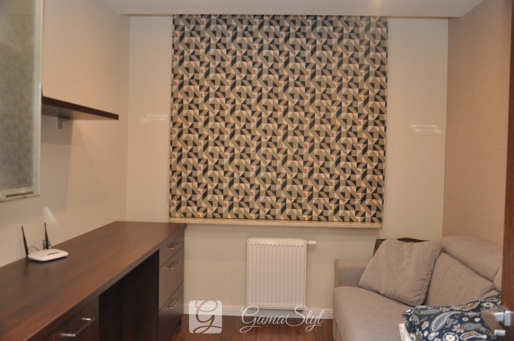 Roleta rzymska z geometrycznym wzorem trójkątów w odcieniach beżu, czerni i czerwieni w pokoju z biurkiem i sofą.