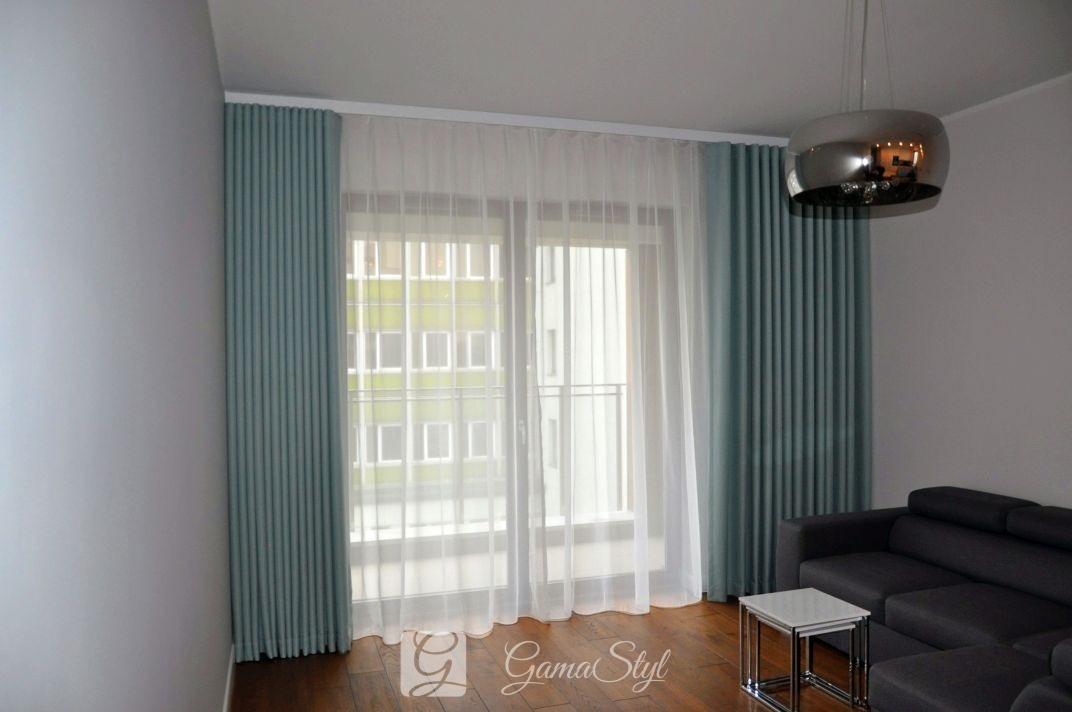 Eleganckie, turkusowe zasłony panelowe z białymi, zwiewnymi firanami w salonie w Warszawie, z widoczną nowoczesną lampą wiszącą i fragmentem szarej sofy.