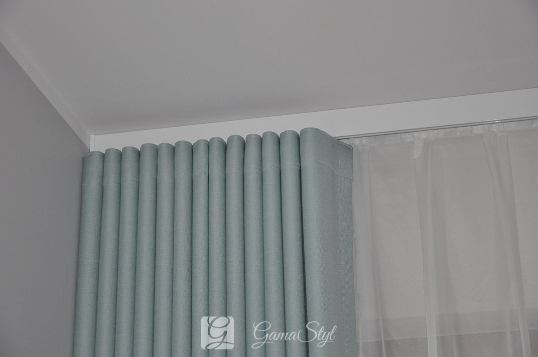 Jasnoniebieska, gęsto marszczona zasłona z podszewką, zawieszona na karniszu, obok firanka z woalu, w jasnym wnętrzu.