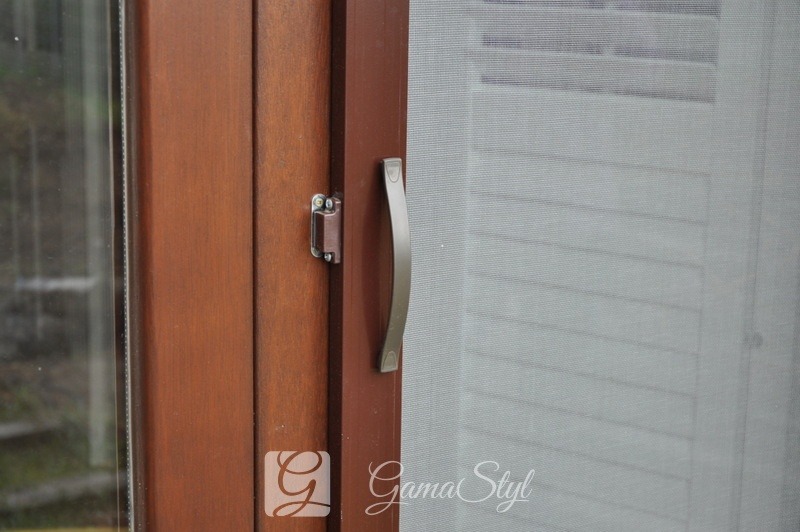 Brązowa rama okienna z zamontowaną plisą w kolorze białym, widoczny uchwyt i zamek okna.