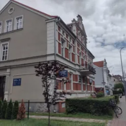 Elewacja budynku kancelarii prawnej z tabliczką informacyjną, widok z ulicy z rowerem opartym o żywopłot.