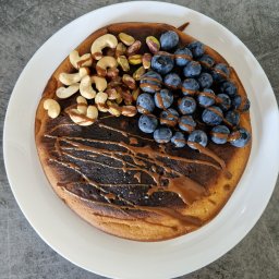 Dietetyka Cattleya - Puszysty naleśnik z polewą karmelową, udekorowany świeżymi borówkami, orzechami nerkowca i pistacjami na białym talerzu.