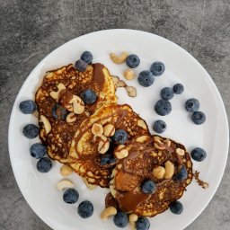 Dietetyka Cattleya - Trzy puszyste naleśniki polane czekoladą, posypane świeżymi borówkami i orzechami, ułożone na białym talerzu na szarym tle.