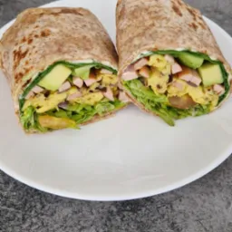 Przekrojony wrap z pełnoziarnistej tortilli z sałatą, jajecznicą, awokado i szynką, ułożony na białym talerzu na szarym tle.