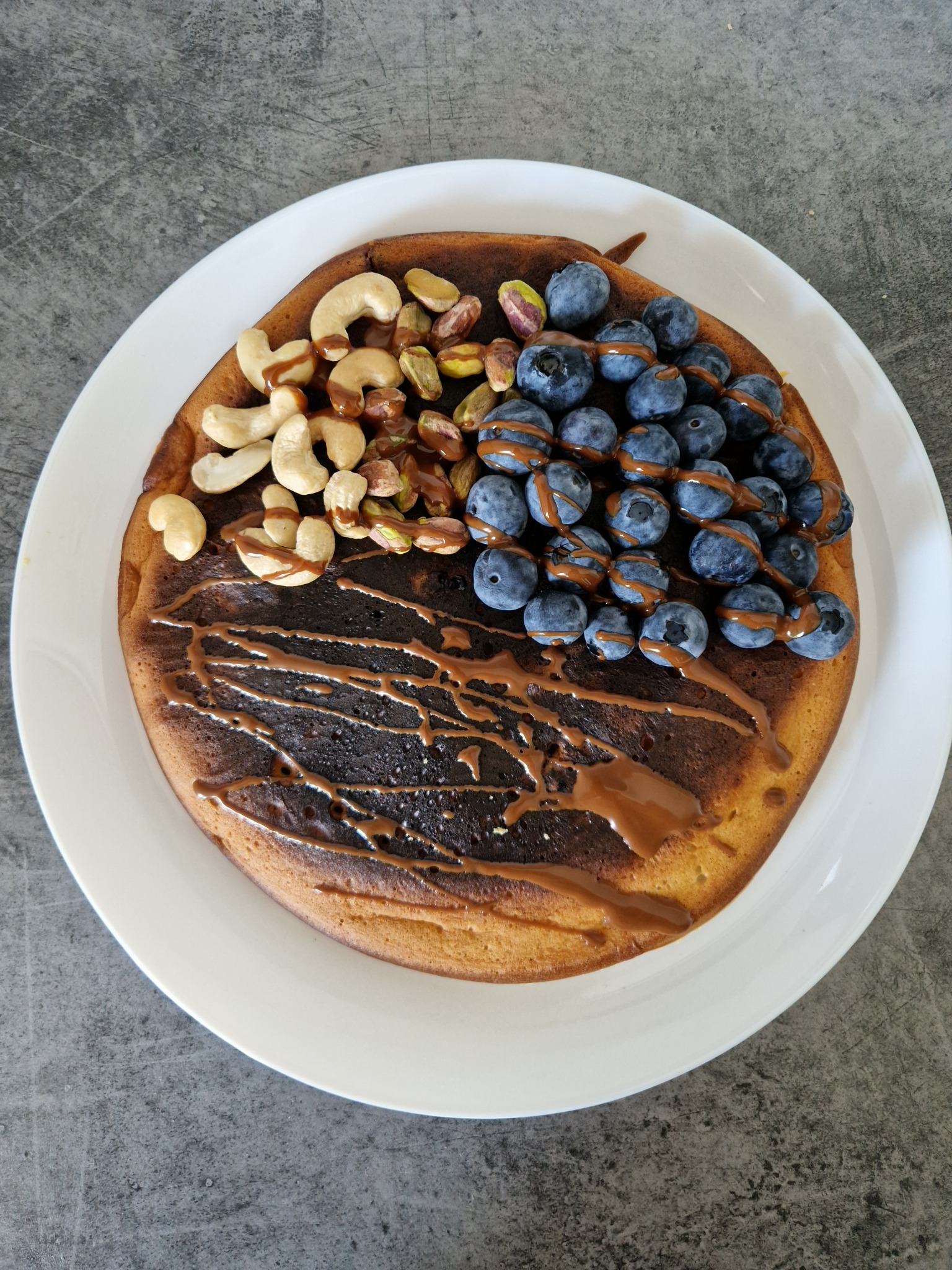 Puszysty naleśnik z polewą karmelową, udekorowany świeżymi borówkami, orzechami nerkowca i pistacjami na białym talerzu.