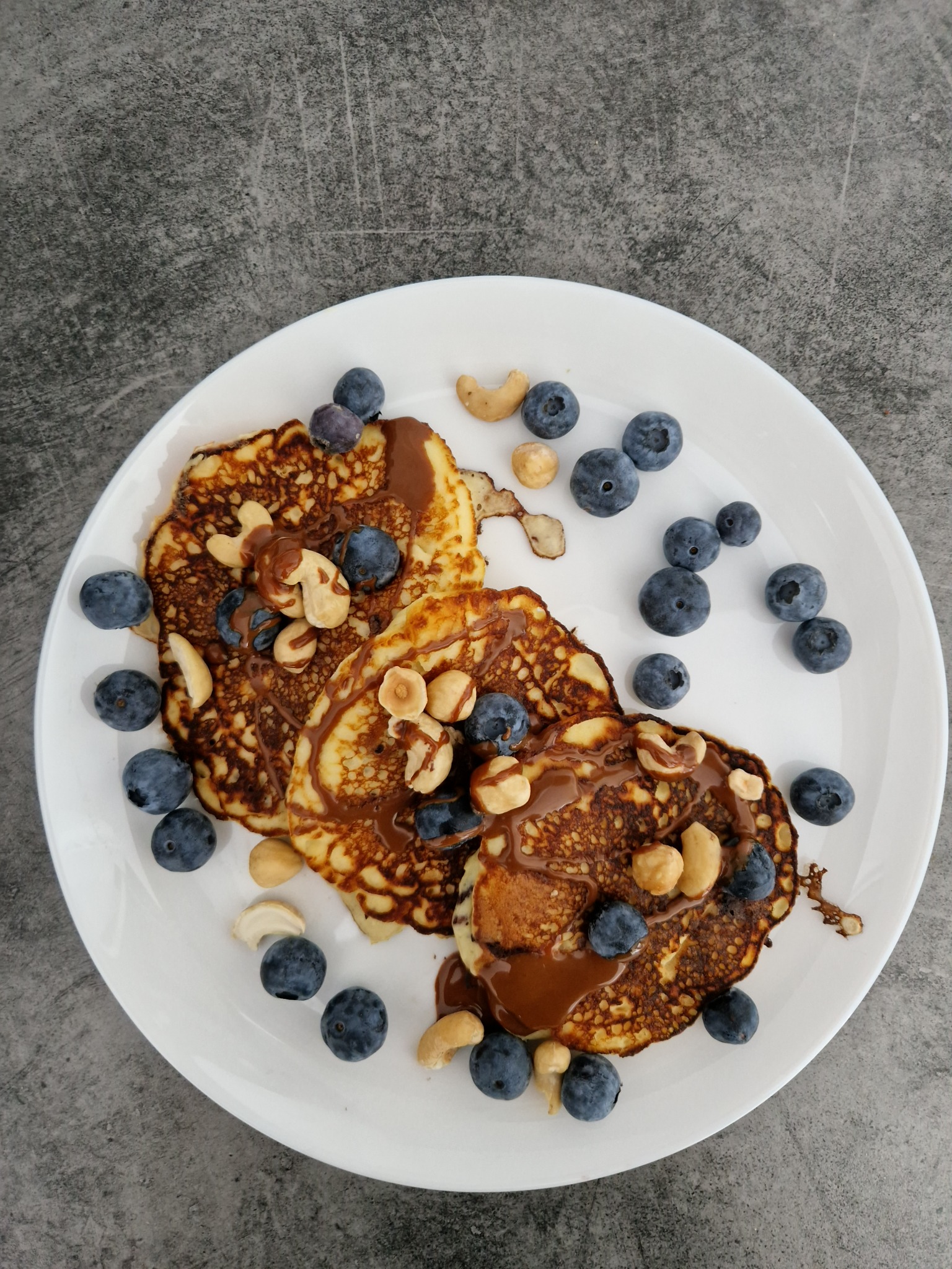 Trzy puszyste naleśniki polane czekoladą, posypane świeżymi borówkami i orzechami, ułożone na białym talerzu na szarym tle.
