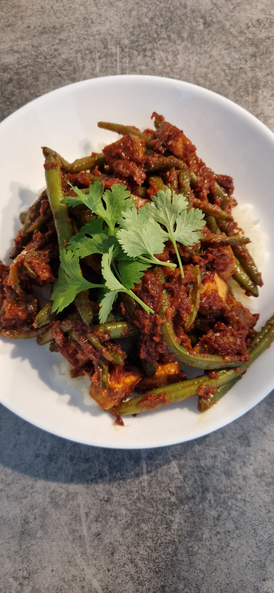 Gotowe danie kuchni azjatyckiej z zielonej fasolki i tofu w sosie, udekorowane świeżą kolendrą, podane na białym ryżu w białej misce.