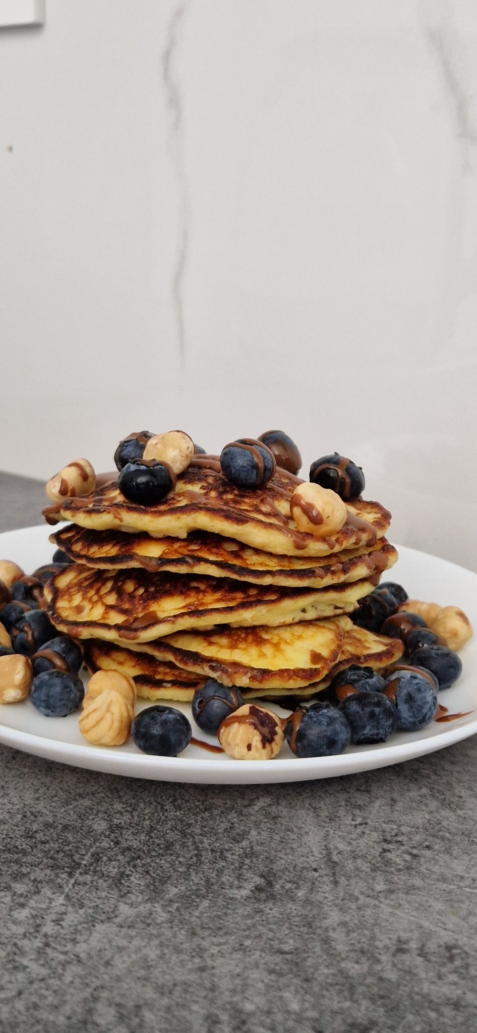 Stos pięciu puszystych naleśników na białym talerzu, polanych sosem czekoladowym i udekorowanych świeżymi borówkami i orzechami laskowymi.