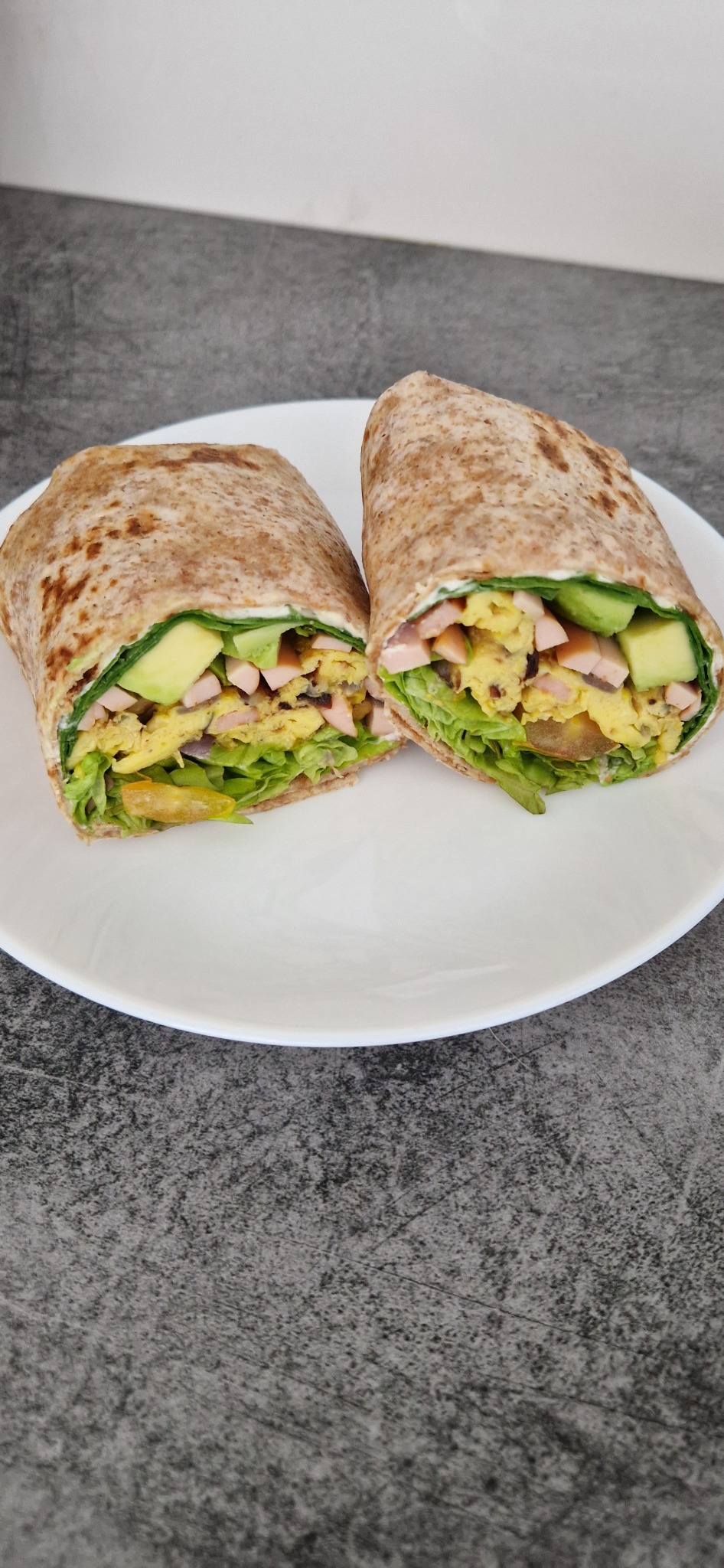 Przekrojony wrap z pełnoziarnistej tortilli z sałatą, jajecznicą, awokado i szynką, ułożony na białym talerzu na szarym tle.