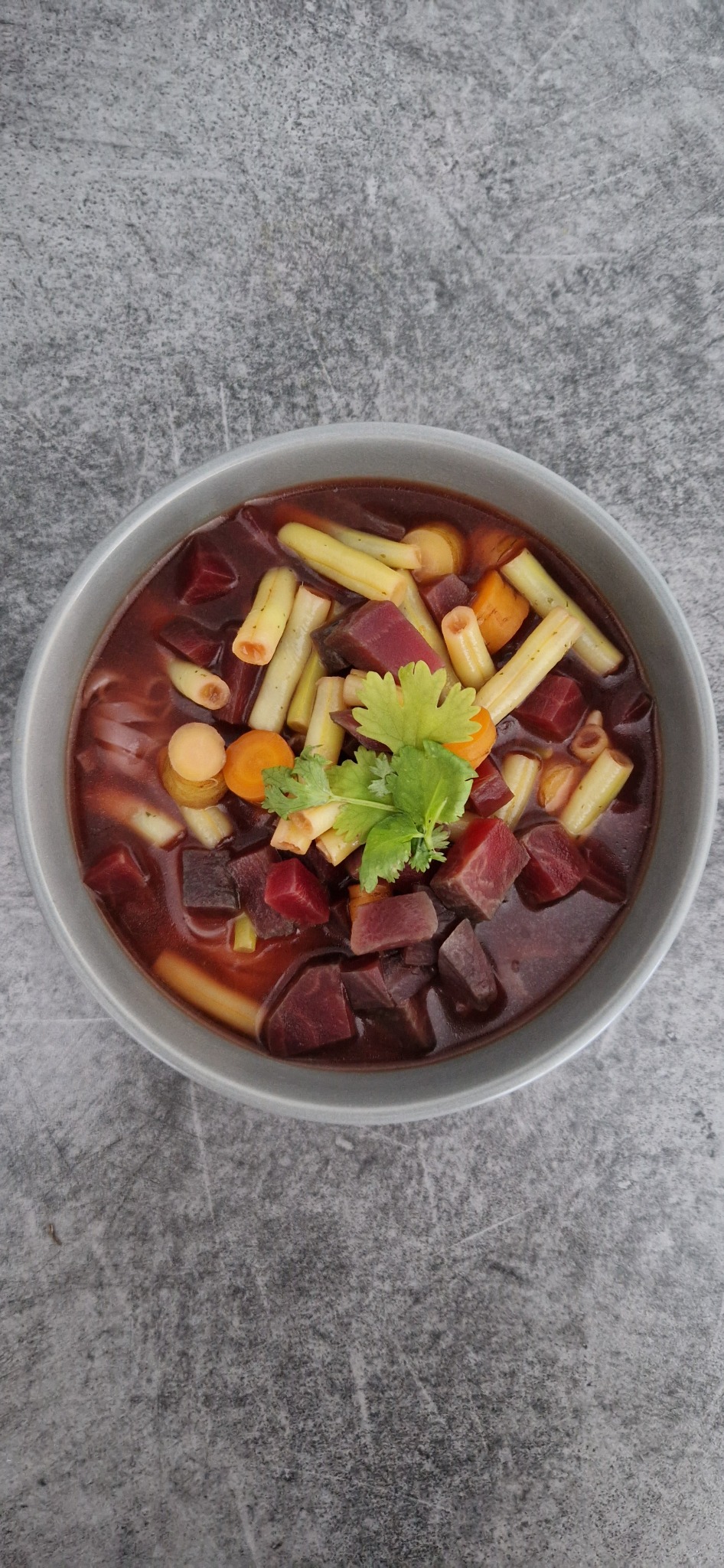 Buraczkowa zupa z makaronem i marchewką, udekorowana zieloną pietruszką, podana w szarej misce na tle szarego blatu.