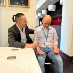 Mężczyzna w garniturze dotyka ręki siedzącego mężczyzny, kt&oacute;ry ma na szyi identyfikator; na stole leżą wizyt&oacute;wki.