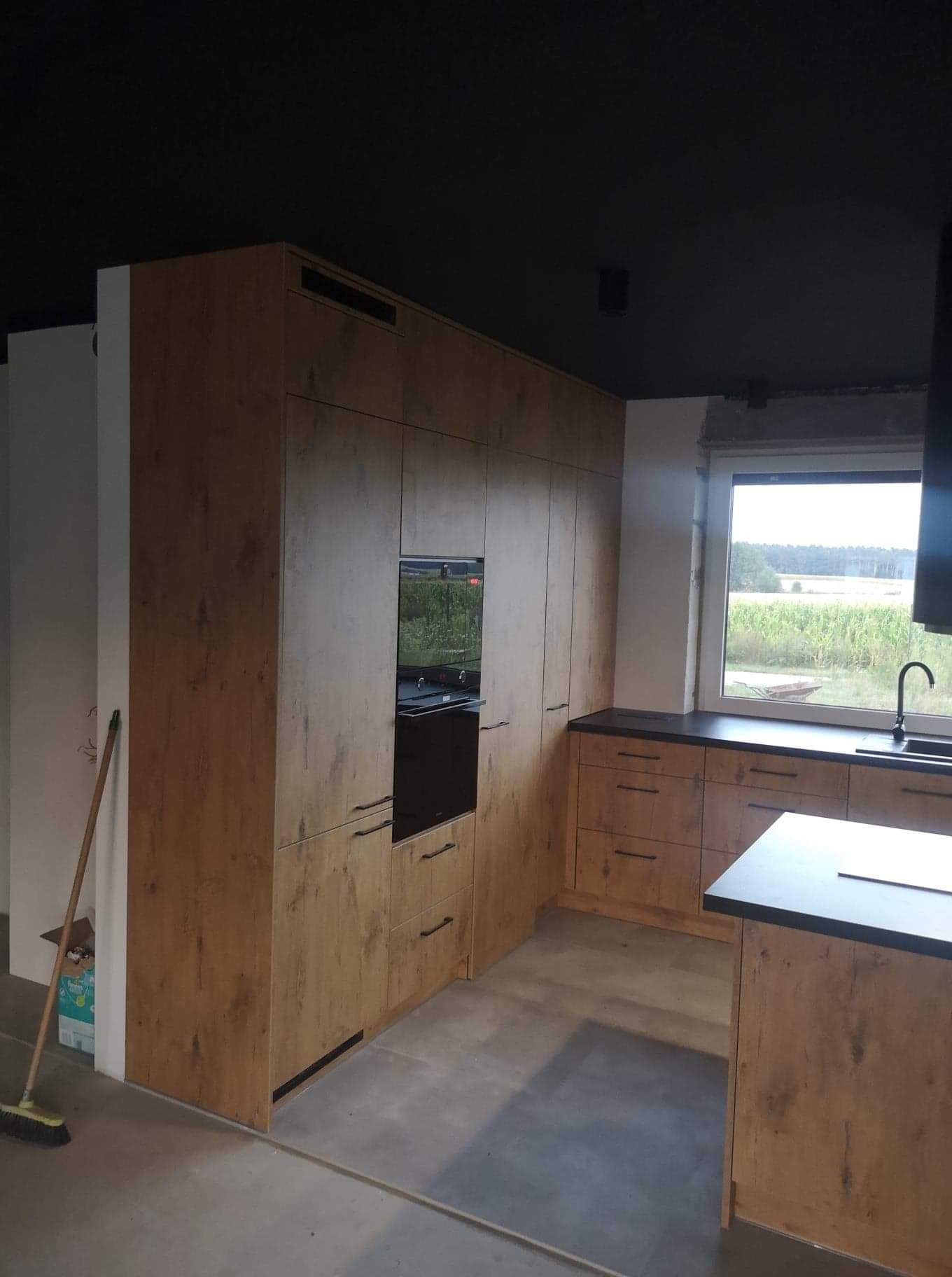 Nowoczesna kuchnia z drewnopodobnymi frontami, czarnym blatem i piekarnikiem w zabudowie, widok na pole za oknem, częściowo posprzątana podłoga.