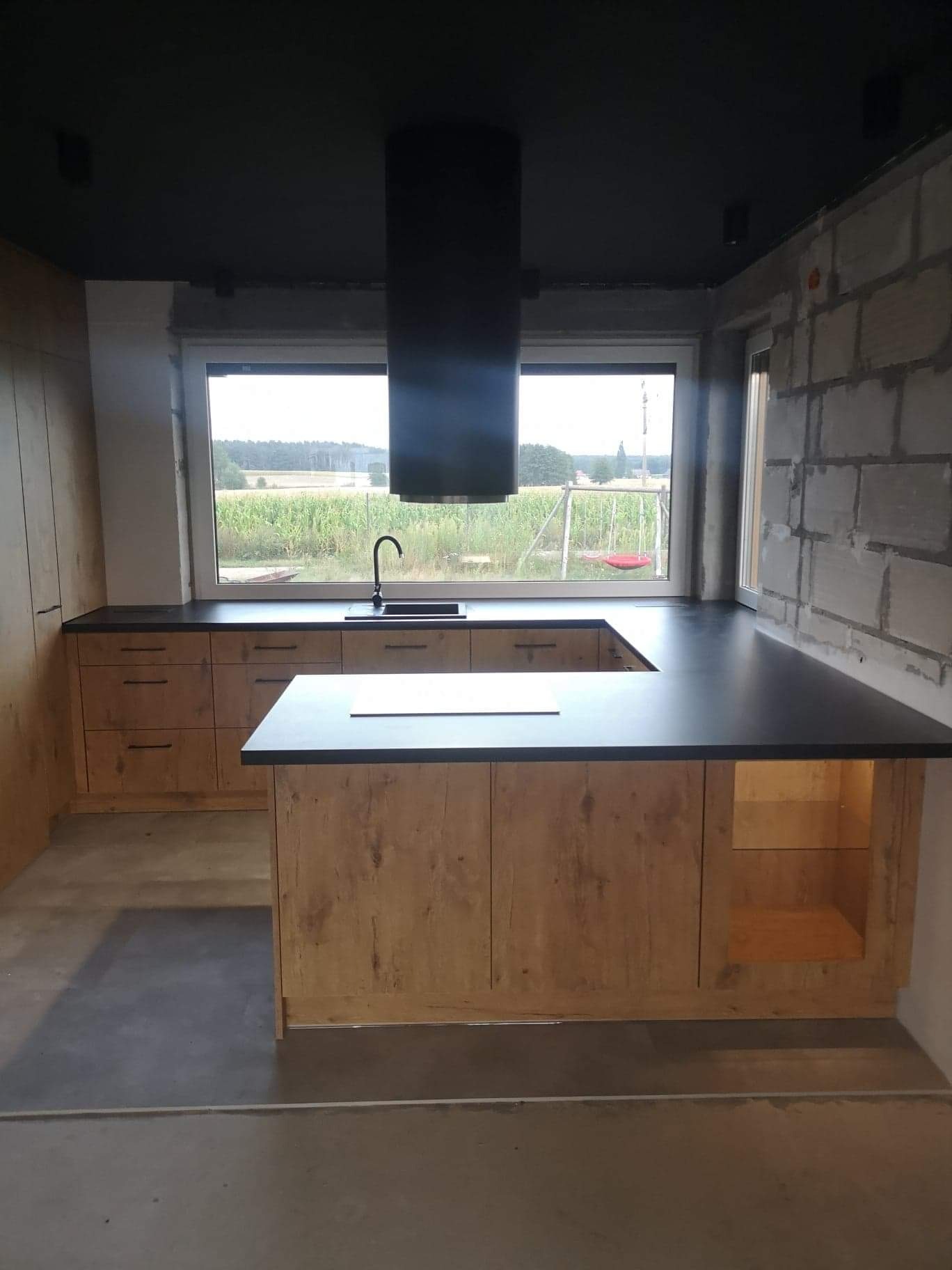 Nowoczesna kuchnia z drewnianymi szafkami i czarnym blatem, widok na pole i huśtawkę przez okno, surowy mur z białego bloczka po prawej.