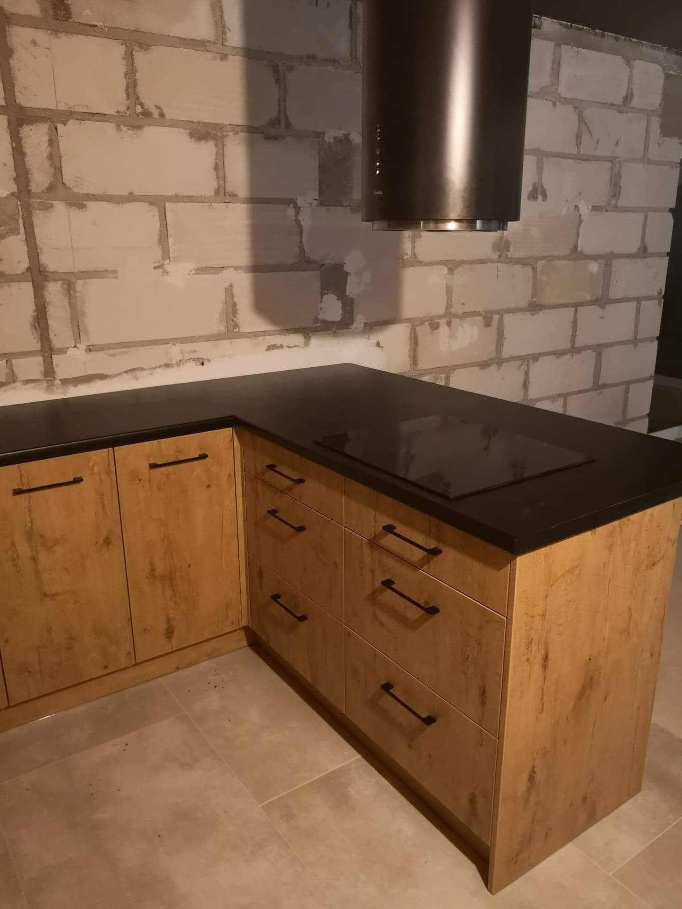 Fragment nowej kuchni z drewnopodobnymi frontami, czarnym blatem i płytą indukcyjną, oraz czarnym okapem na tle surowej ściany z bloczków.