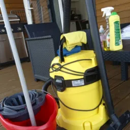 Żółty odkurzacz uniwersalny Kärcher WD na drewnianym tarasie obok czerwonego wiadra Vileda z mopem, butelki ze środkami czyszczącymi i mebli ogrodowych.