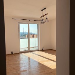 Aka-bud POLSKIE BUDOWNICTWO - Jasne, przestronne wnętrze z drewnopodobną podłogą, dużym oknem balkonowym z widokiem na miasto i nowoczesną lampą wiszącą.