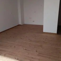 Puste, jasne pomieszczenie z drewnianą podłogą, świeżo po położeniu, widoczne gniazdka elektryczne na ścianie.