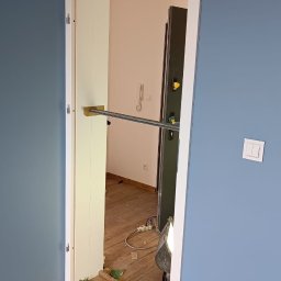 Aka-bud POLSKIE BUDOWNICTWO - Montaż ościeżnicy drzwiowej w Lęborku, widoczne niebieskie ściany, fragment nowej podłogi i lampa budowlana oświetlająca wnętrze.