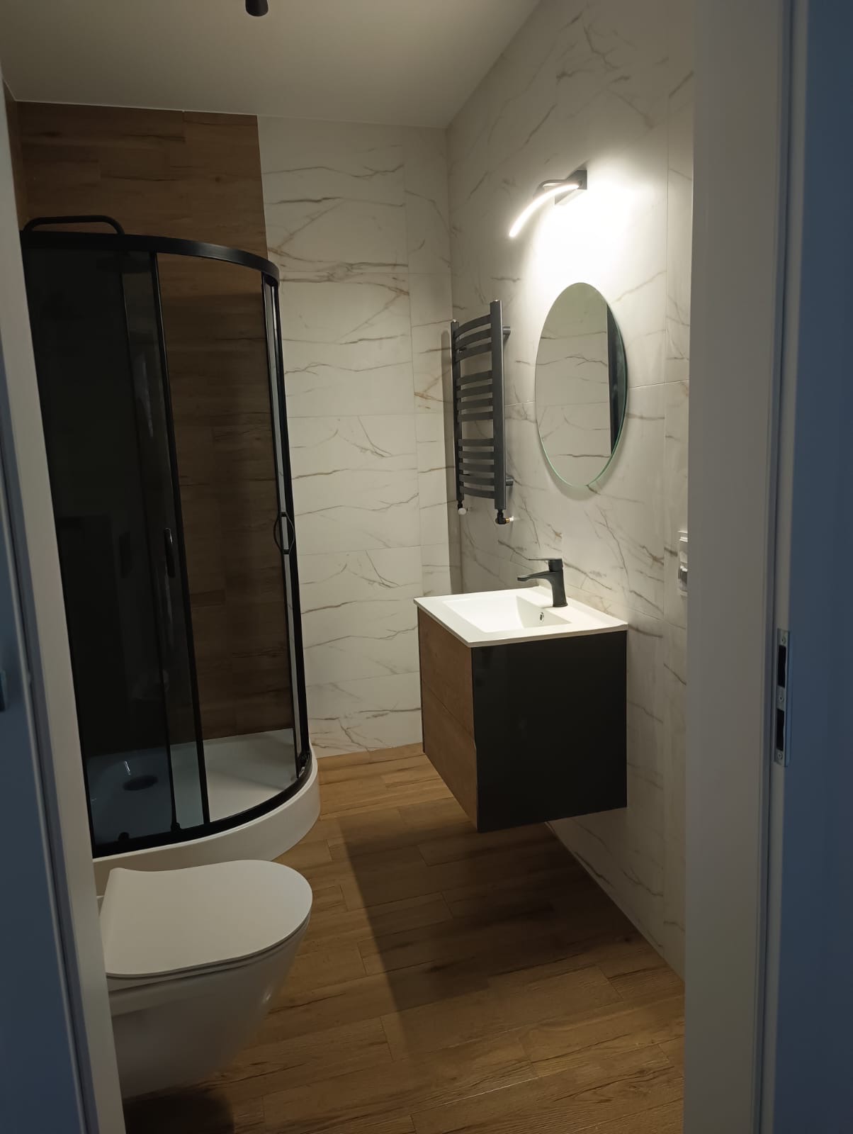 Nowoczesna łazienka z kabiną prysznicową z czarnymi profilami, umywalką nablatową z szafką w kolorze drewna i czerni, okrągłym lustrem podświetlanym LED i grzejnikiem drabinkowym.