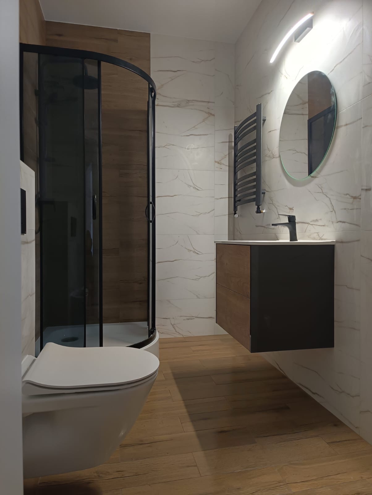 Nowoczesna łazienka z kabiną prysznicową w czarnej ramie, wiszącą szafką z umywalką, okrągłym lustrem z podświetleniem i grzejnikiem drabinkowym na ścianie wyłożonej płytkami imitującymi marmur...