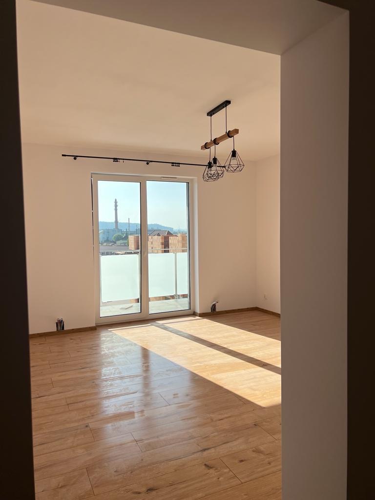 Jasne, przestronne wnętrze z drewnopodobną podłogą, dużym oknem balkonowym z widokiem na miasto i nowoczesną lampą wiszącą.
