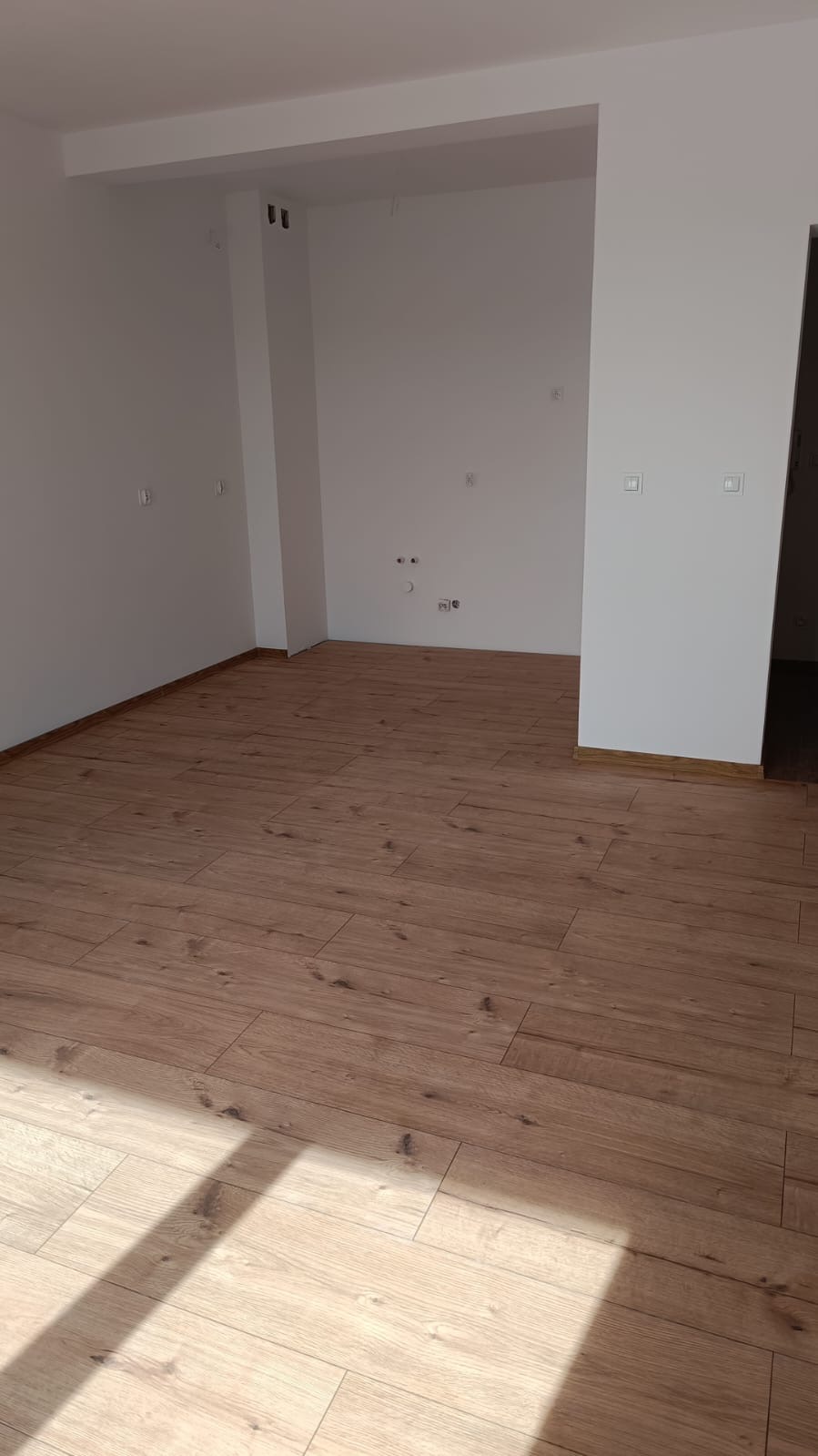 Puste, jasne pomieszczenie z drewnianą podłogą, świeżo po położeniu, widoczne gniazdka elektryczne na ścianie.