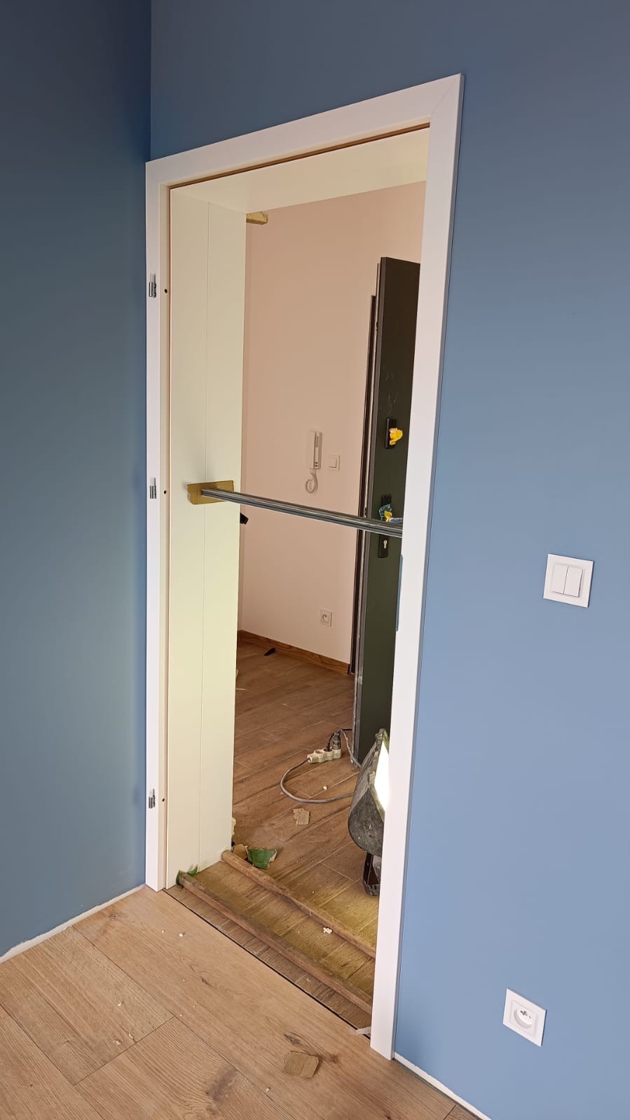 Montaż ościeżnicy drzwiowej w Lęborku, widoczne niebieskie ściany, fragment nowej podłogi i lampa budowlana oświetlająca wnętrze.