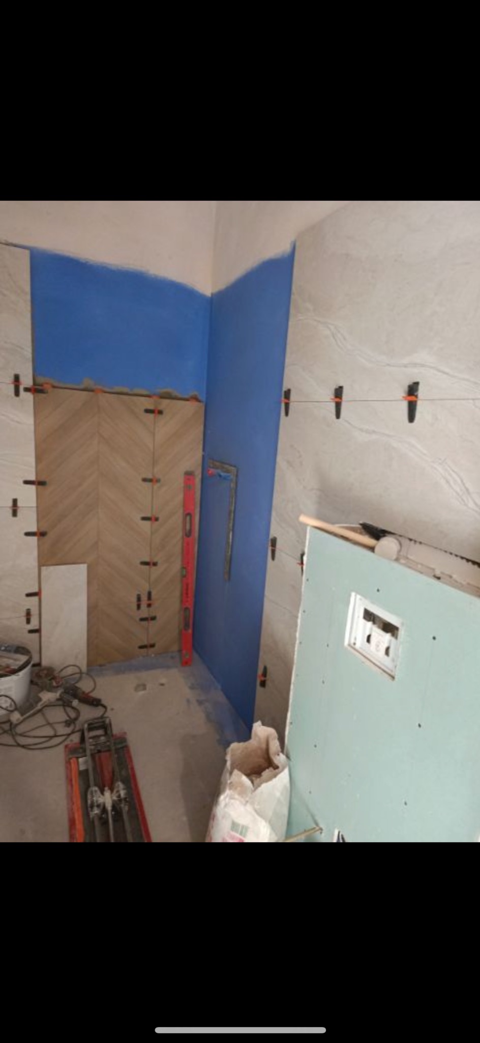 Wnętrze łazienki w trakcie prac: częściowo ułożone płytki w jodełkę, wyrównane poziomnicą, niebieska farba na ścianie, płyta gipsowo-kartonowa z otworem na rewizję, narzędzia glazurnicze na podłodze.