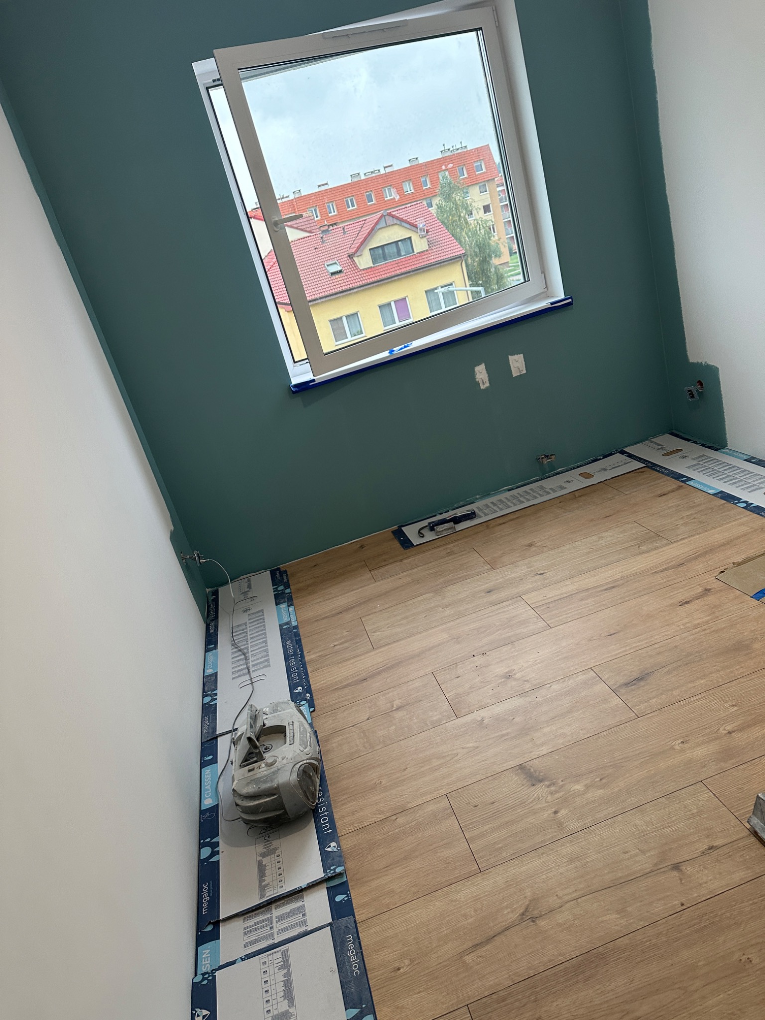 Pokój w trakcie odświeżania: ściana pomalowana na zielono w górnej części, biała w dolnej, drewniana podłoga częściowo zakryta kartonami ochronnymi, widoczne gniazdka elektryczne i okno z widokiem...
