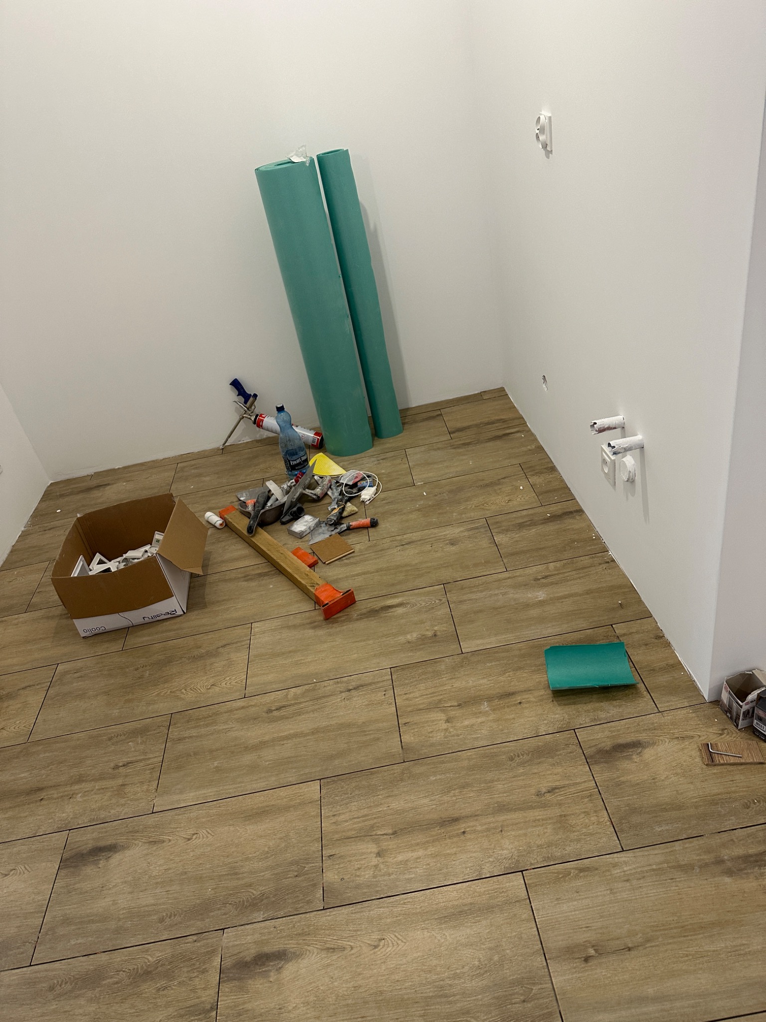 Podłoga w trakcie układania płytek imitujących drewno, widoczne narzędzia glazurnicze, rolki materiału izolacyjnego i karton z elementami wykończeniowymi w rogu pomieszczenia.