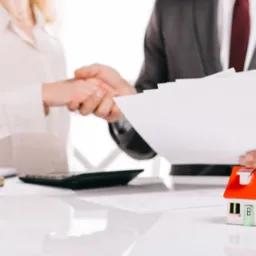 Uścisk dłoni nad stołem z kalkulatorem, pieczątką i modelem domu z czerwoną dachówką, mężczyzna w garniturze trzyma plik dokumentów.