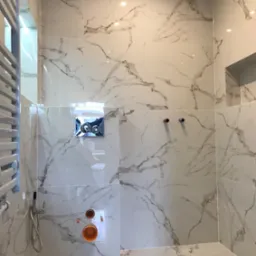 Ściana i podłoga w łazience wyłożone białymi płytkami imitującymi marmur, widoczne elementy instalacji wodnej i grzejnik drabinkowy, stan surowy przed montażem armatury.