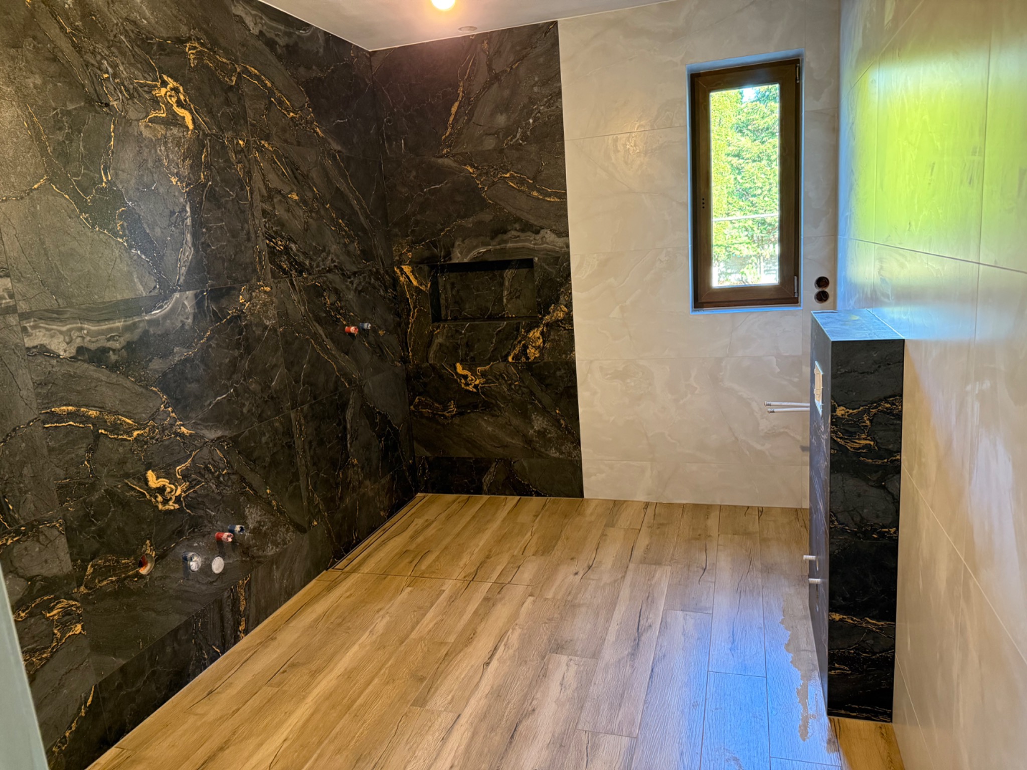 Nowoczesna łazienka w trakcie wykończenia: ściana z czarnego marmuru ze złotymi żyłami, jasne płytki imitujące marmur, drewnopodobne panele na podłodze, wnęka w ścianie, okno z widokiem na zieleń.