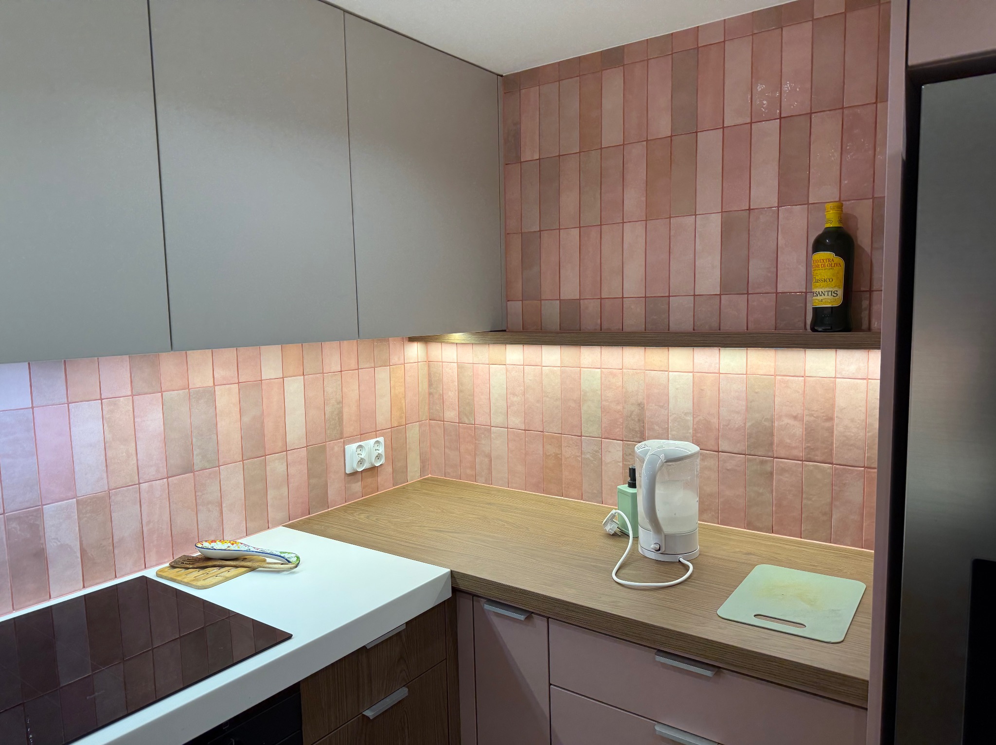 Kuchnia po remoncie z różowymi, pionowo ułożonymi płytkami na ścianie, drewnianym blatem i szarymi szafkami górnymi. Na blacie czajnik elektryczny, deska do krojenia i butelka oliwy.
