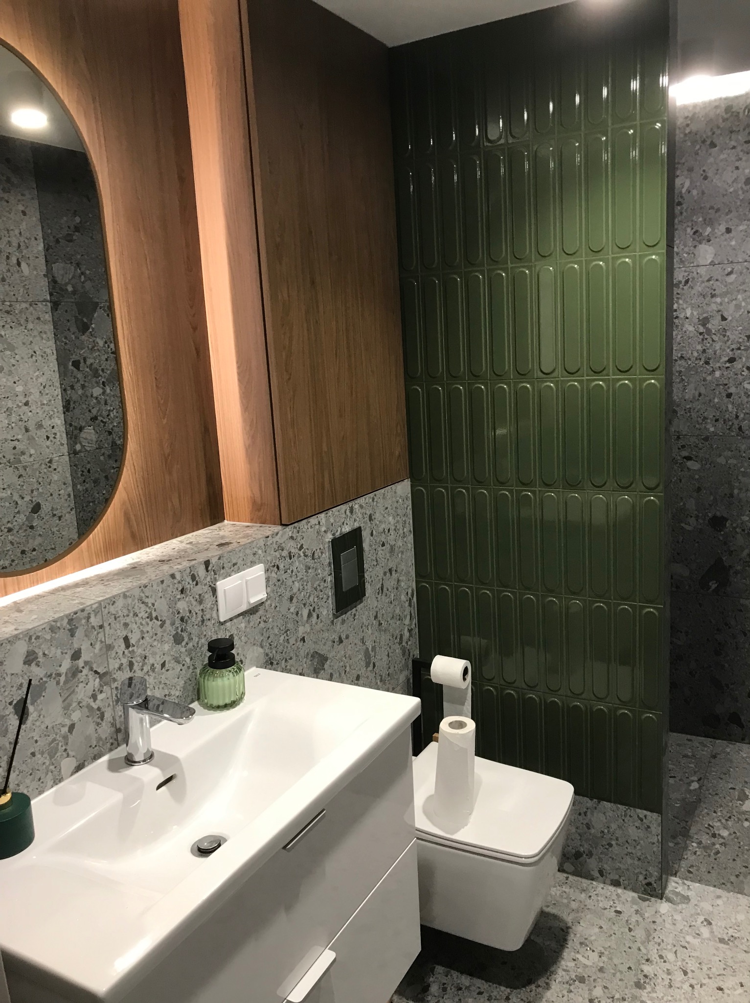 Nowoczesna łazienka z umywalką nablatową, toaletą wiszącą i zielonymi, podłużnymi płytkami na ścianie, uzupełniona drewnianą szafką i lustrem.