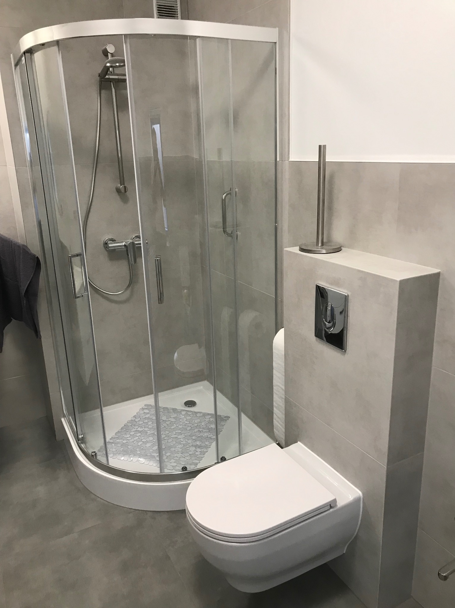 Nowoczesna łazienka z kabiną prysznicową narożną i podwieszaną toaletą w szarej kolorystyce, widoczna mata antypoślizgowa w kabinie.