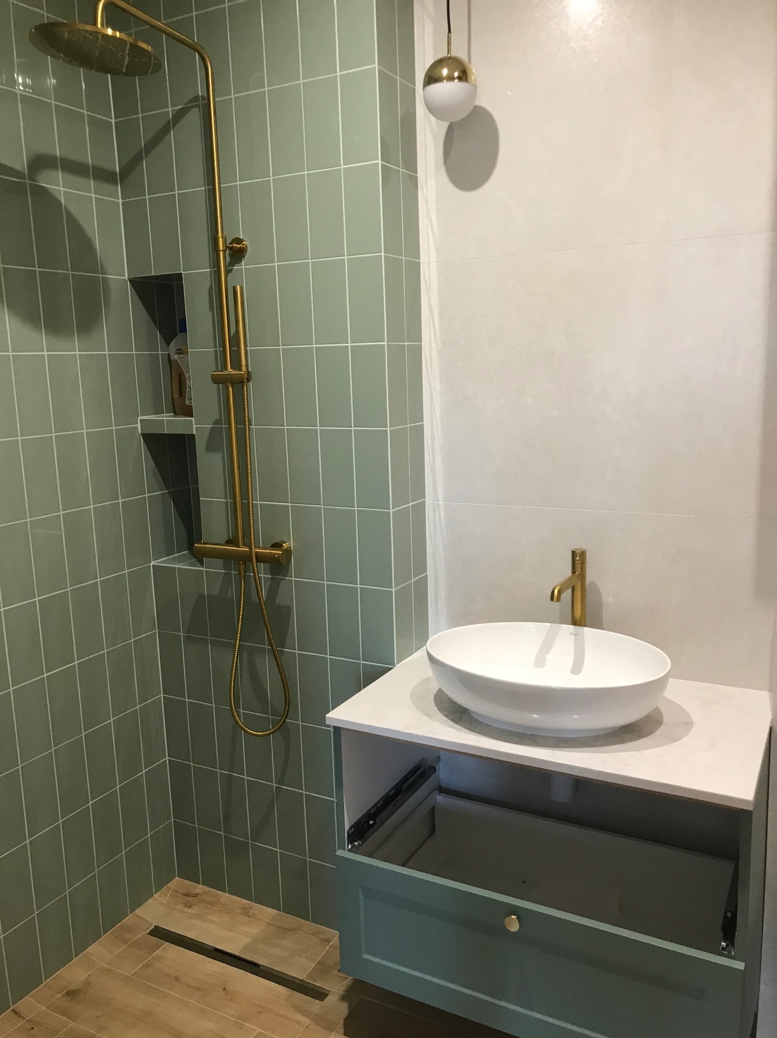 Nowoczesna łazienka z zielonymi płytkami, złotą armaturą, białą umywalką nablatową i szafką z otwartą szufladą.