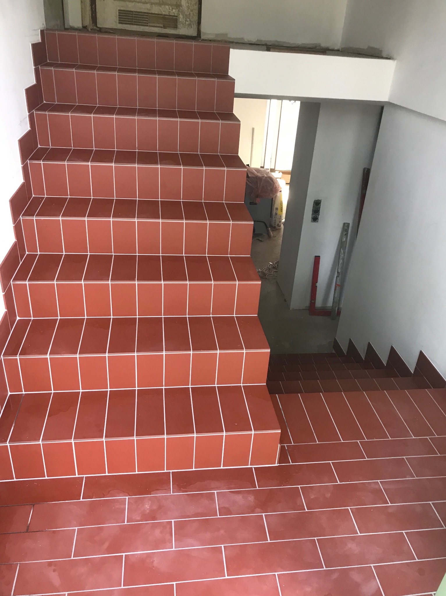 Schody wyłożone terakotą w kolorze ceglastym z jasną fugą, widok z góry, w tle widoczne materiały budowlane i poziomica.