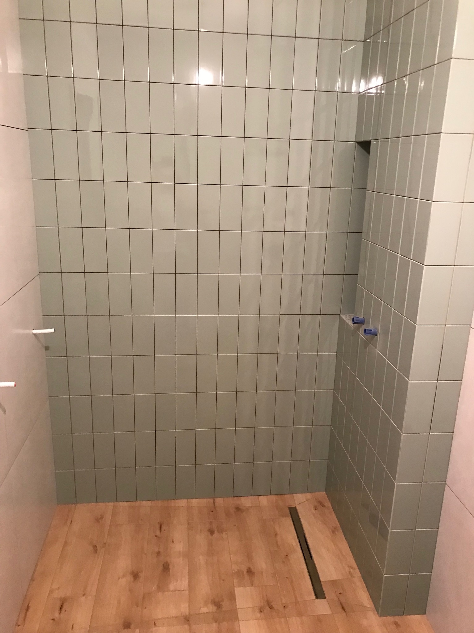 Świeżo wykafelkowany prysznic z odpływem liniowym i drewnopodobną podłogą, widoczne wystające rurki do podłączenia armatury.