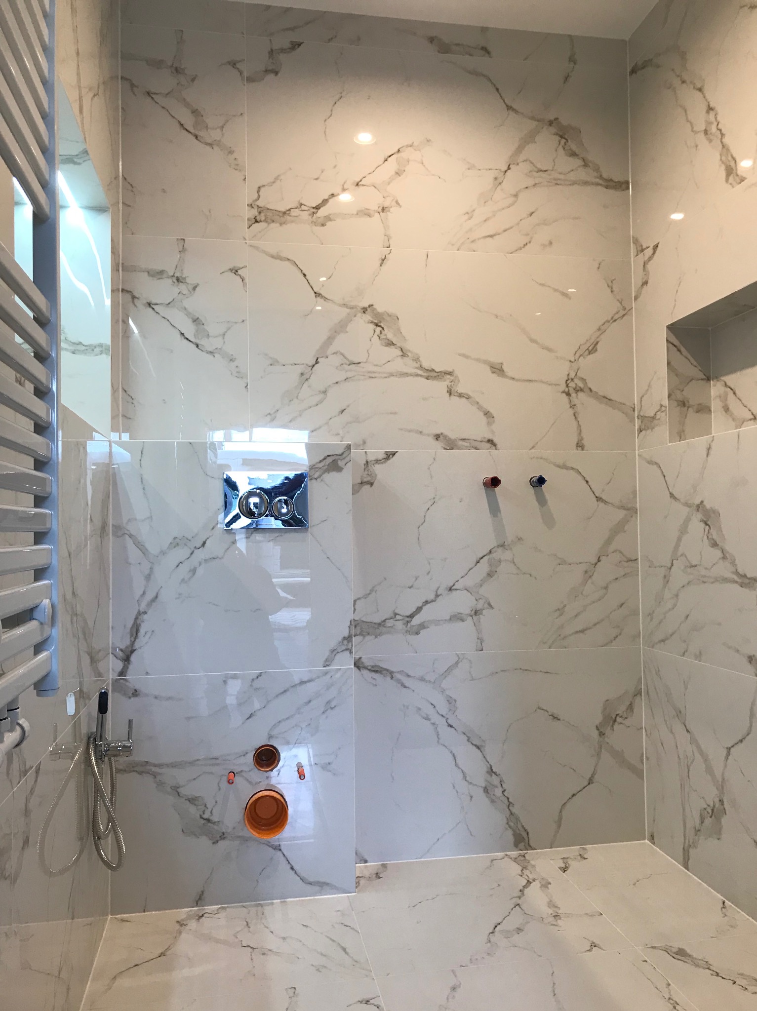 Ściana i podłoga w łazience wyłożone białymi płytkami imitującymi marmur, widoczne elementy instalacji wodnej i grzejnik drabinkowy, stan surowy przed montażem armatury.