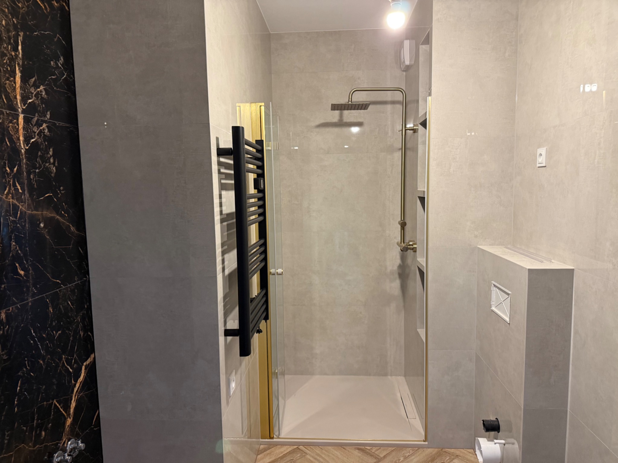 Nowoczesna łazienka z kabiną prysznicową typu walk-in, grzejnikiem drabinkowym i płytkami imitującymi marmur. Złote wykończenia armatury i ramy kabiny.