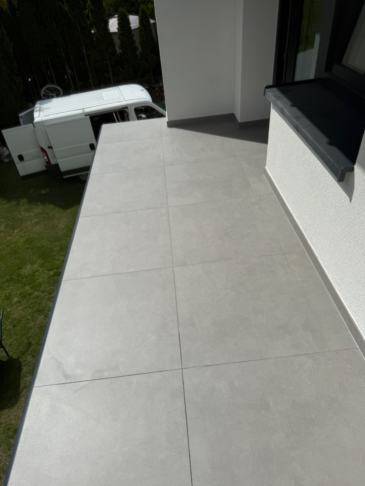 Jasnoszara, kwadratowa terakota na balkonie z widokiem na trawnik i zaparkowany biały samochód dostawczy z otwartymi drzwiami.