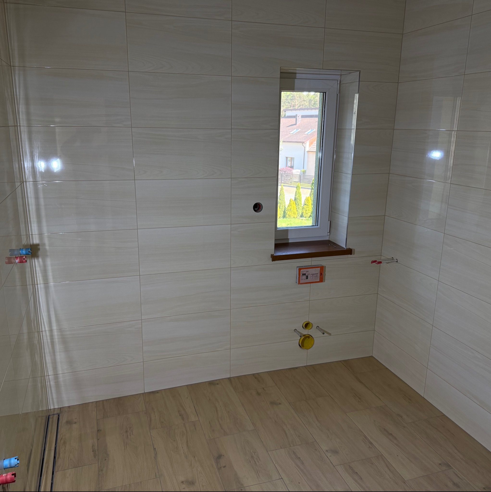 Jasna łazienka po remoncie: beżowe płytki na ścianach i podłodze imitujące drewno, okno z widokiem na zieleń, przygotowane instalacje wod-kan.