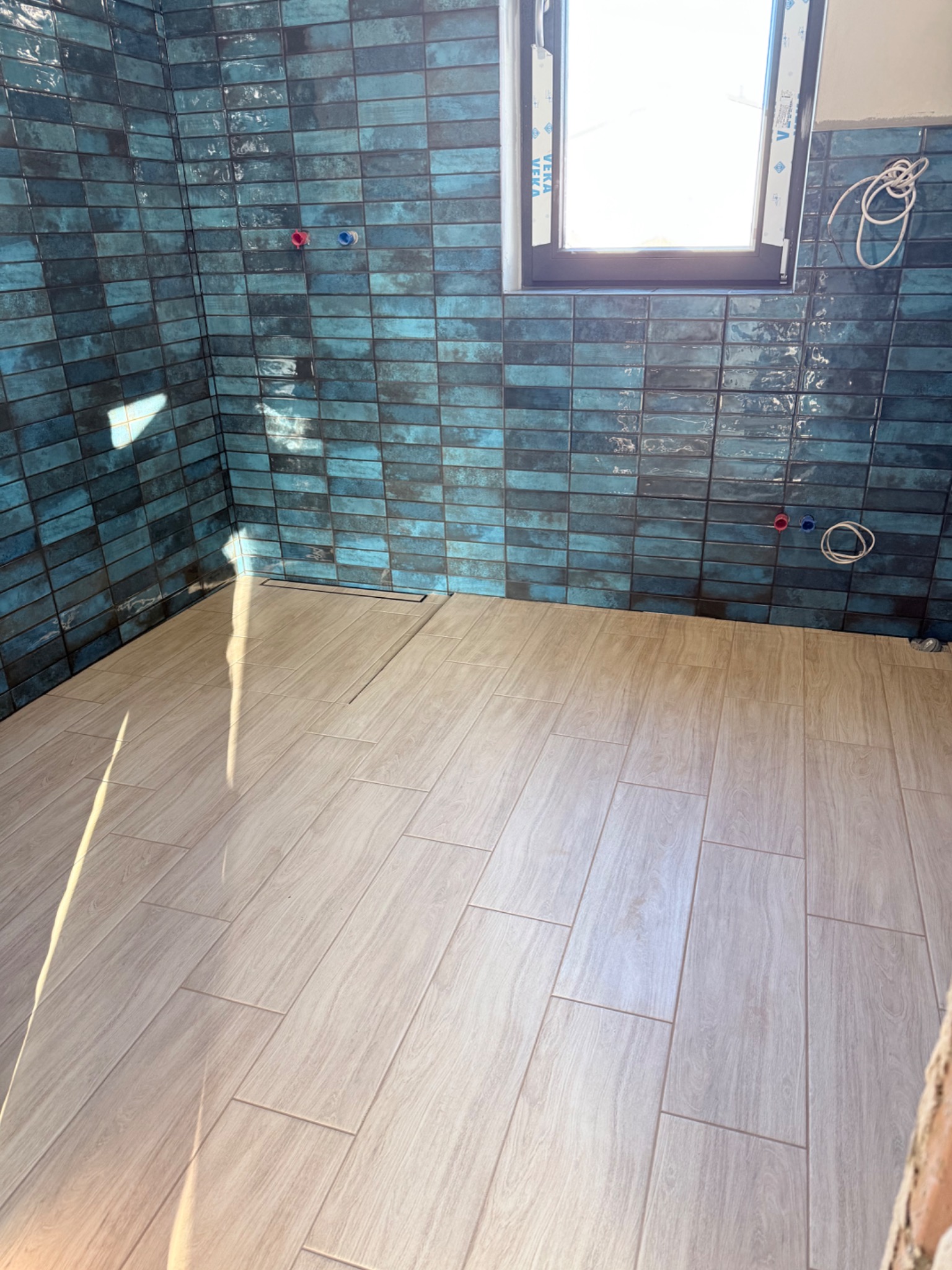 Wykończona łazienka z turkusową glazurą ułożoną w cegiełkę i jasnymi panelami podłogowymi imitującymi drewno. Widoczne przyłącza wodne i okno.