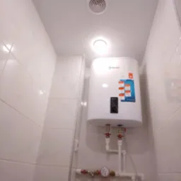 Biały bojler elektryczny marki Thermex, zamontowany na ścianie wyłożonej białymi płytkami w łazience, widoczne rury doprowadzające wodę i liczniki zużycia, oświetlenie sufitowe.