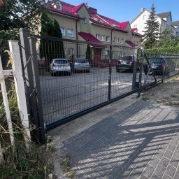 Konsta Sp.zo.o - Czarna brama wjazdowa panelowa z widokiem na parking przed budynkiem z czerwonym dachem w słoneczny dzień.