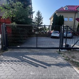 Konsta Sp.zo.o - Zewnętrzna brama przesuwna z panelowym wypełnieniem, widoczna z zewnątrz posesji, z szarej kostki brukowej, w tle dwa zaparkowane samochody i budynki mieszkalne w słoneczny dzień.