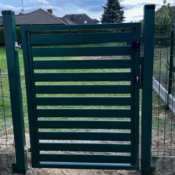 Nowoczesna, zielona furtka z poziomych paneli, wkomponowana w ogrodzenie z siatki, na tle trawnika i domu z dach&oacute;wką.