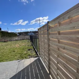 Nowoczesne ogrodzenie drewniane z poziomych desek, widok z boku na tle trawnika, domu i błękitnego nieba z chmurami.